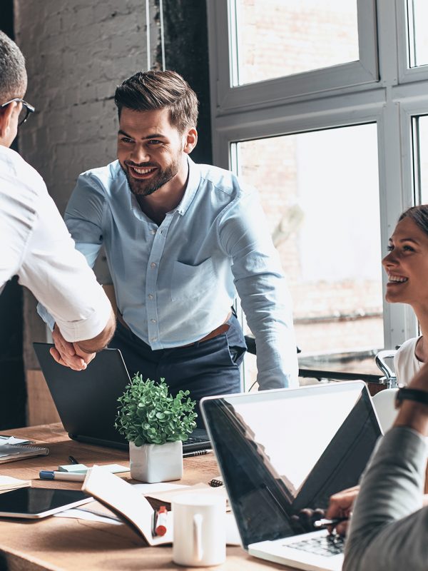 Young modern men in smart casual wear shaking hands and smiling while working in the creative office